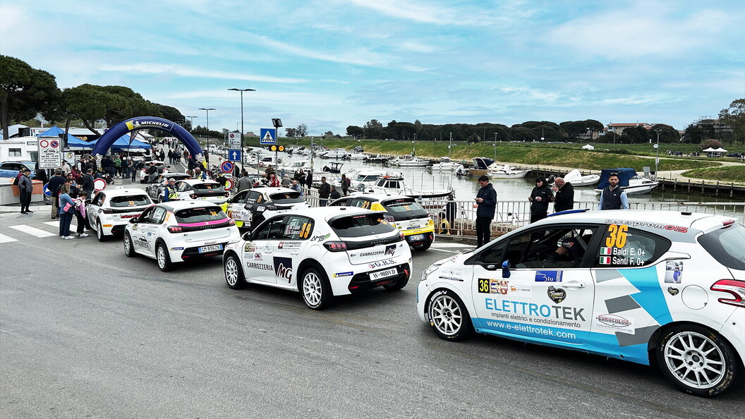 Una fila di auto da rally bianche in coda su una strada vicino a un porto, pronte per la partenza del rally Trofeo Maremma. Spettatori e barche ormeggiate sono visibili.
