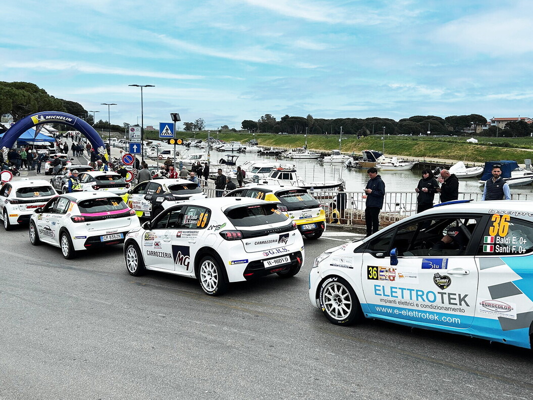 Una fila di auto da rally bianche in coda su una strada vicino a un porto, pronte per la partenza del rally Trofeo Maremma. Spettatori e barche ormeggiate sono visibili.