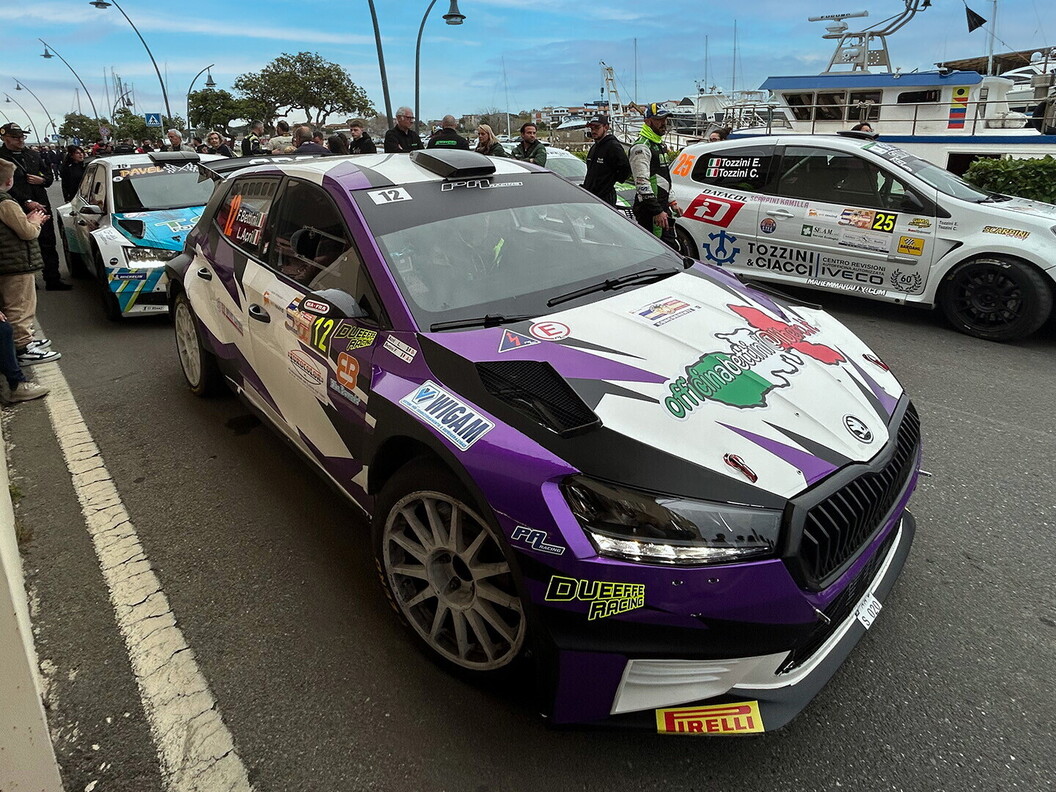 Un'auto viola da rally con la livrea della squadra è ferma in una strada affollata vicino a un porto, con spettatori e un'altra auto sullo sfondo.