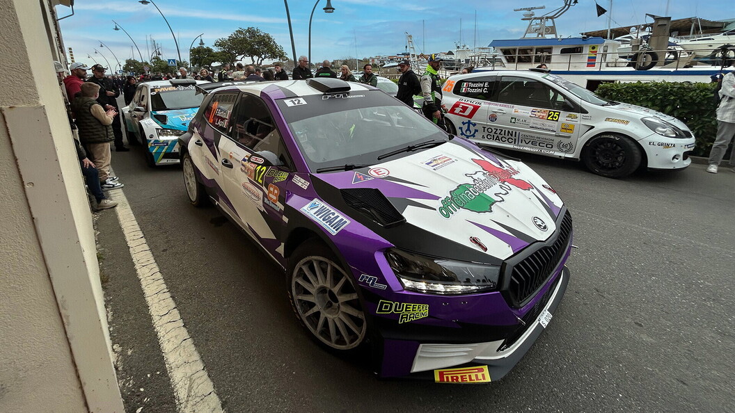 Un'auto viola da rally con la livrea della squadra è ferma in una strada affollata vicino a un porto, con spettatori e un'altra auto sullo sfondo.