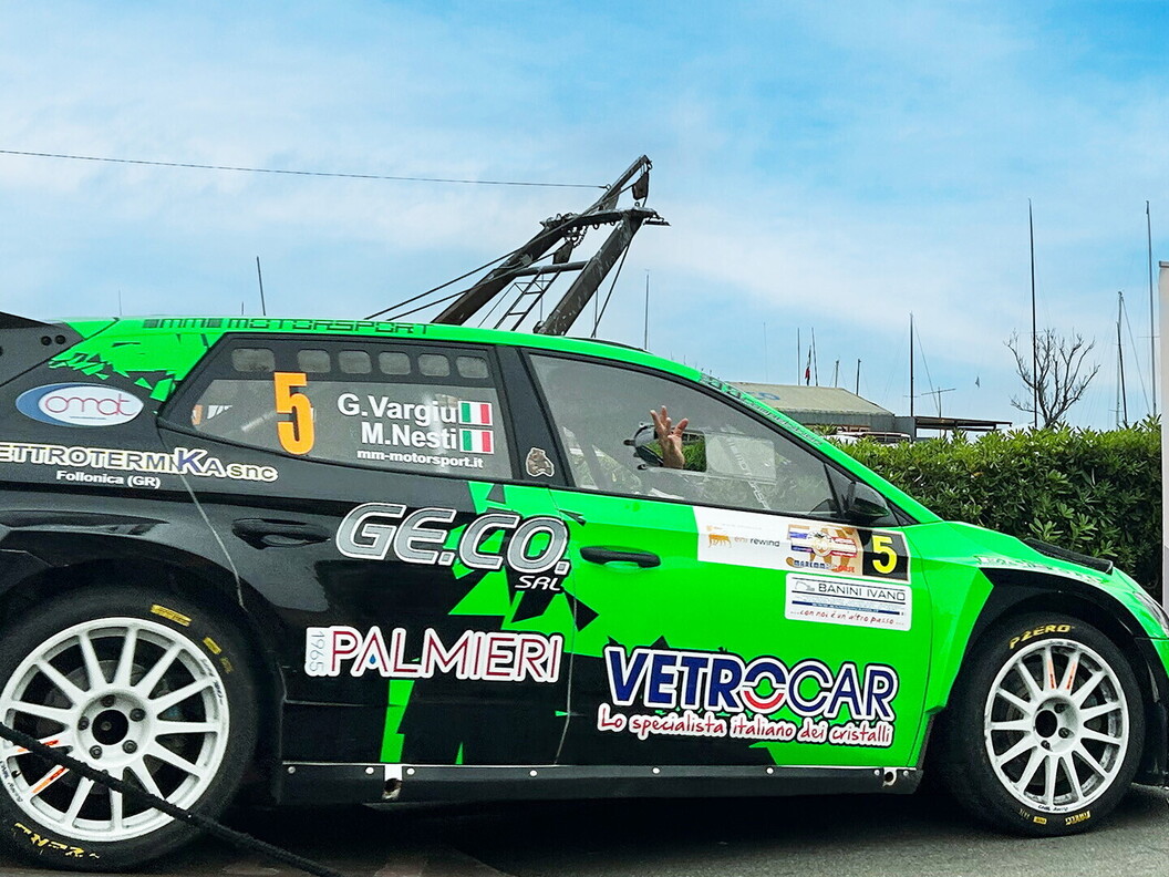L'auto numero 5 di Vargiu e Nesti, una Skoda verde brillante con sponsor Palmieri e Vetrocar, parcheggiata davanti allo striscione Eni Rewind. Un membro del team saluta dall'abitacolo.