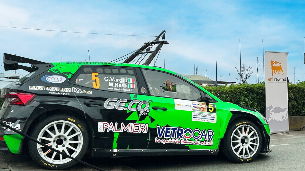 L'auto numero 5 di Vargiu e Nesti, una Skoda verde brillante con sponsor Palmieri e Vetrocar, parcheggiata davanti allo striscione Eni Rewind. Un membro del team saluta dall'abitacolo.