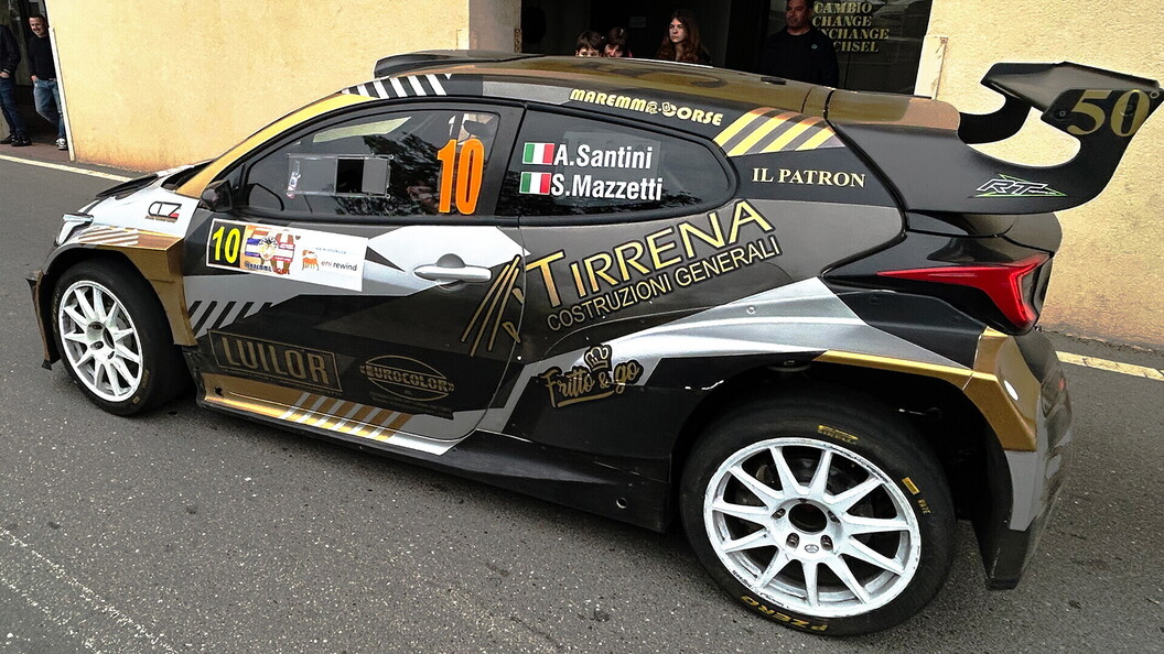 Rally car number 10 with a black and gold livery parked in front of a historic building with spectators nearby.