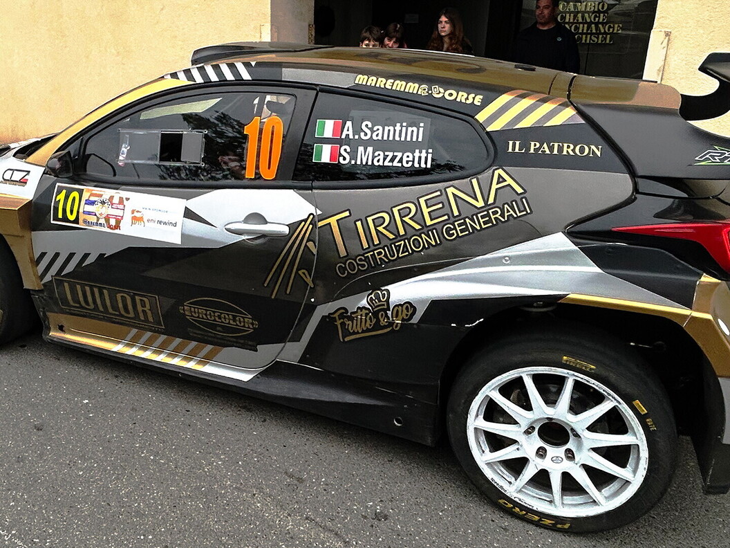 Rally car number 10 with a black and gold livery parked in front of a historic building with spectators nearby.