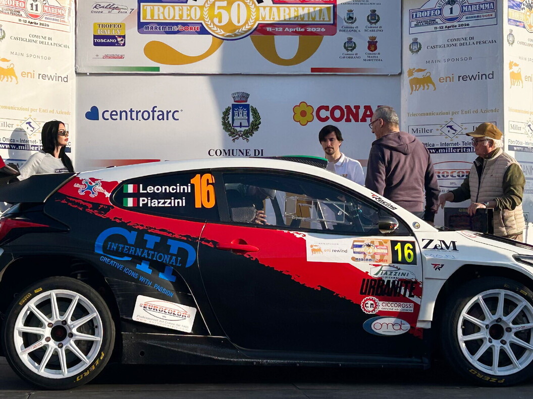 Rally car number 16 with a red, white, and black livery positioned in front of the official event backdrop and sponsors.